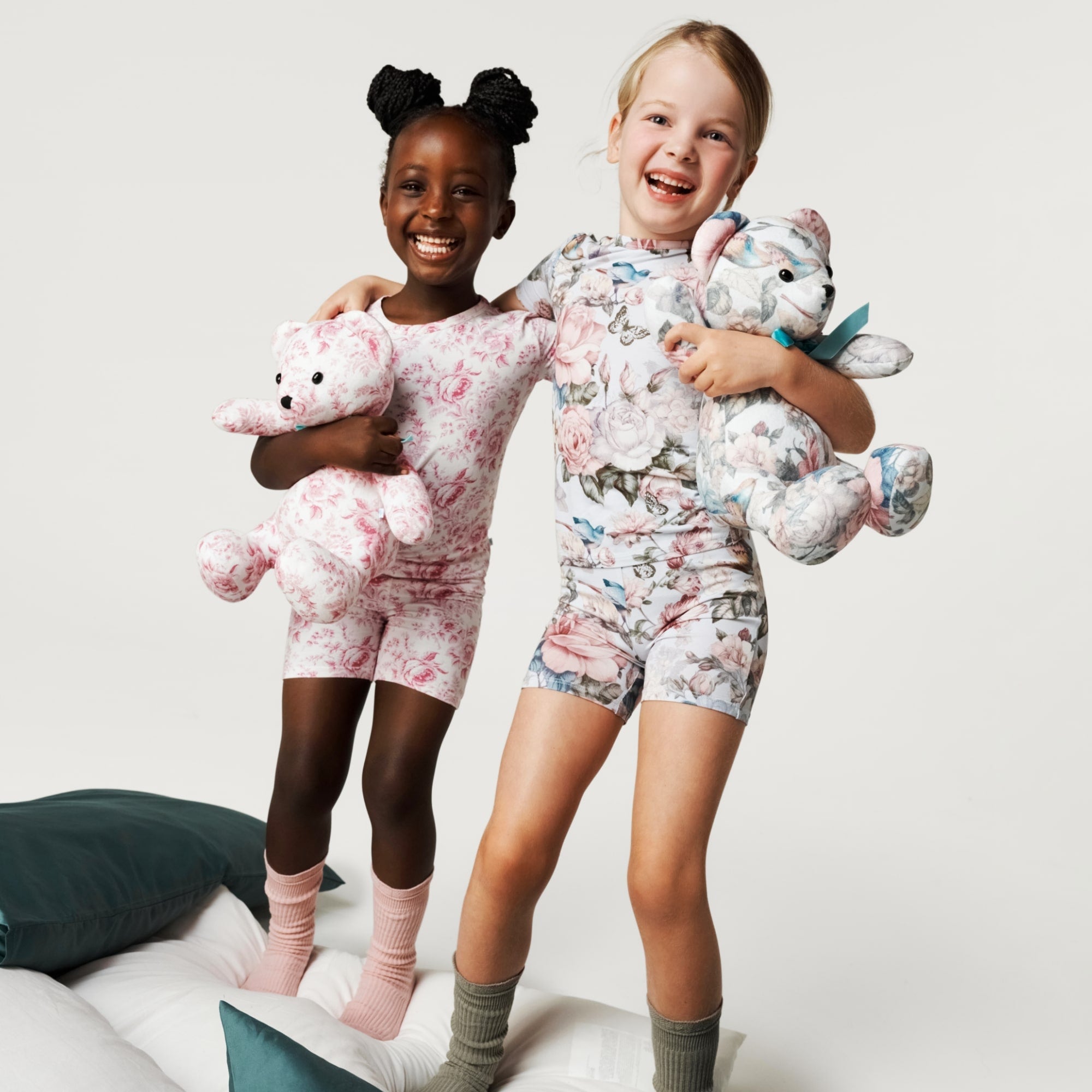 two young girls wearing floral print pajamas holding matching stuffed bears while jumping on bed
